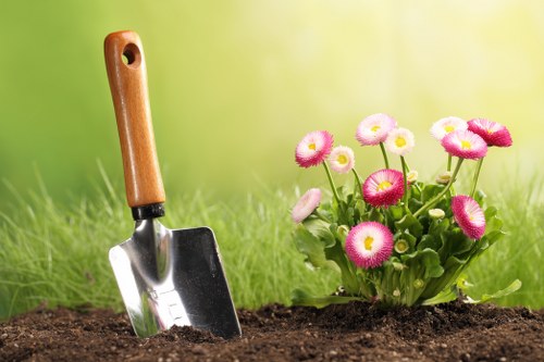 A close-up view of a garden with a freshly dug patch of rich, dark soil in the foreground, featuring a small, stainless steel trowel with a wooden handle inserted into the soil. To the right, there is a vibrant cluster of pink and white flowering plants, possibly primroses, with rounded petals and green foliage, set against a softly blurred background of bright green grass and a subtle gradient of natural daylight. The scene depicts a well-maintained outdoor garden space, suitable for lawn care or planting activities, typical of residential landscaping in Richmond or surrounding areas, highlighting the natural textures and colors common in UK gardens. The image emphasizes garden planting and maintenance elements without showing any additional tools or structures, aligning with professional gardening services like those offered by Lawn Mowing Richmond.