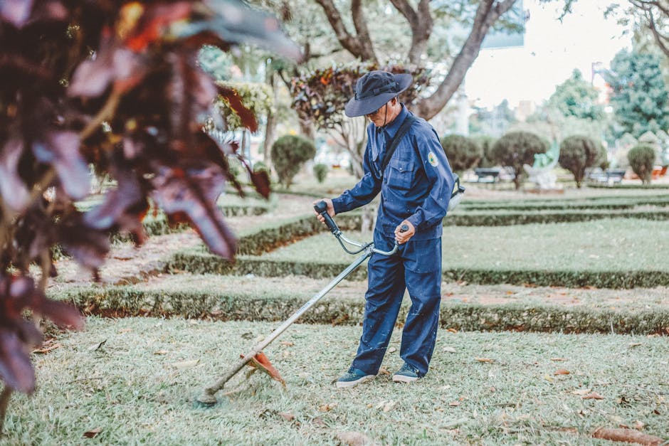 A gardener dressed in a blue uniform and wide-brimmed hat is operating a string trimmer to trim the grass in a well-maintained park or garden. The scene features neatly edged lawn areas, bordered by t