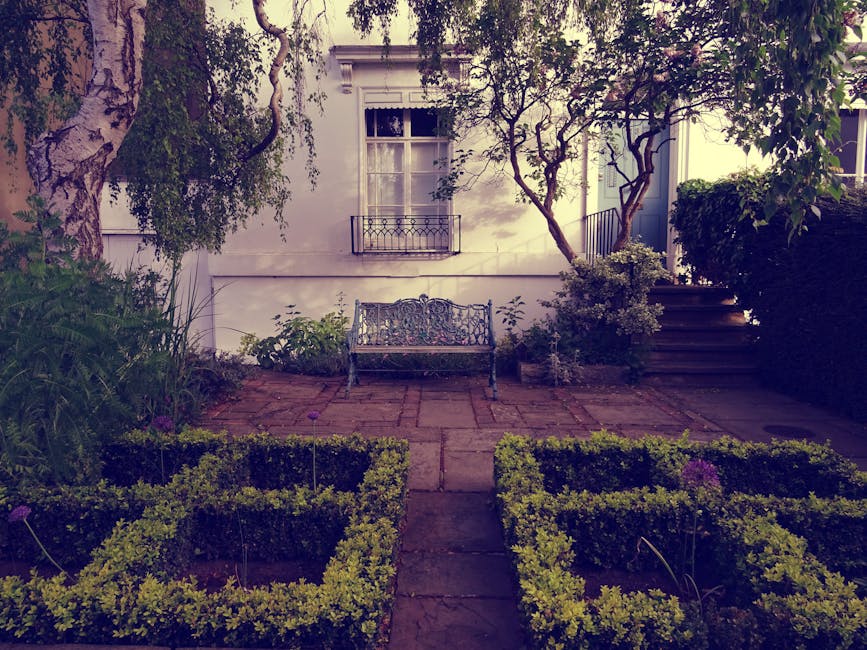 A small, well-maintained garden area in front of a white residential building featuring a central paved pathway leading to a decorative wrought iron bench positioned against the building's wall. The g